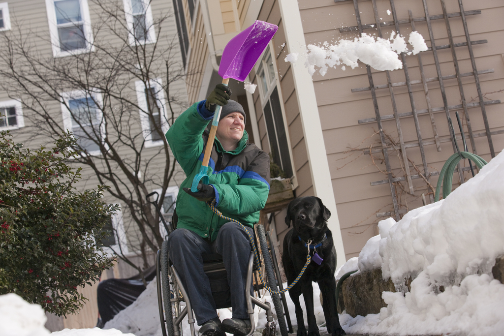 Photo of a person in a wheel chair with a service dog shoveling snow from a sidewalk