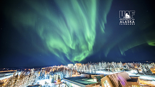The aurora lights up the winter night sky over the Wood Center with the UAF logo in the upper right corner.
